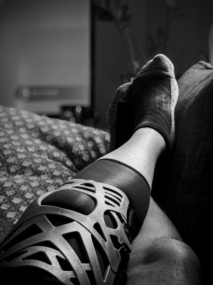 Monochrome photo of a leg wearing a knee brace, resting on a couch.