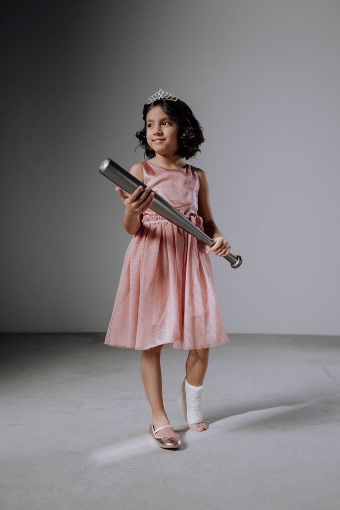 A young girl with a leg cast holding a baseball bat, wearing a pink dress and tiara, standing confidently.