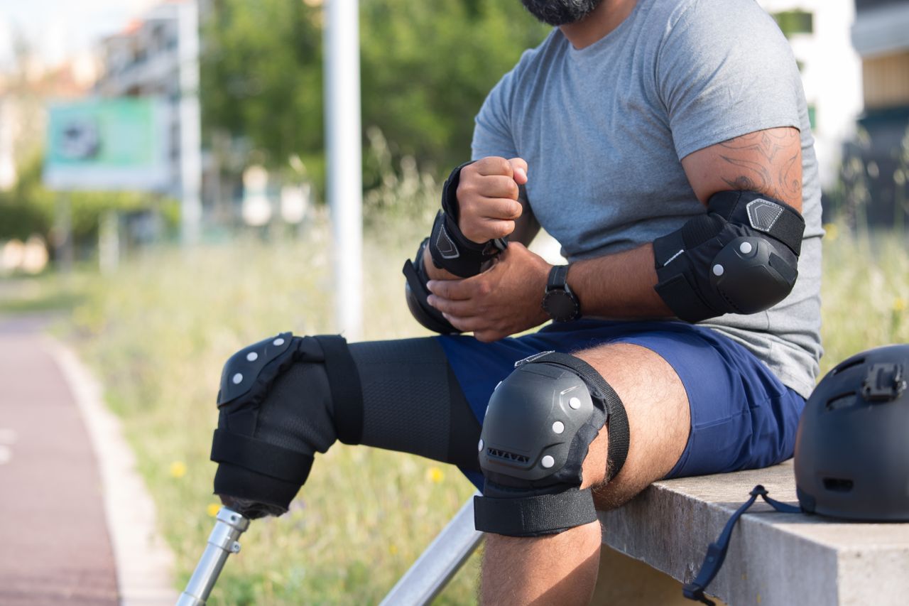 Man with prosthetic leg wearing protective gear, preparing for skateboarding outdoors.